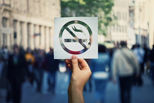 Hand holding a paper sheet with no smoking sign over a crowded street public place background. Forbidden area zone, restrictive symbol stop smoke.